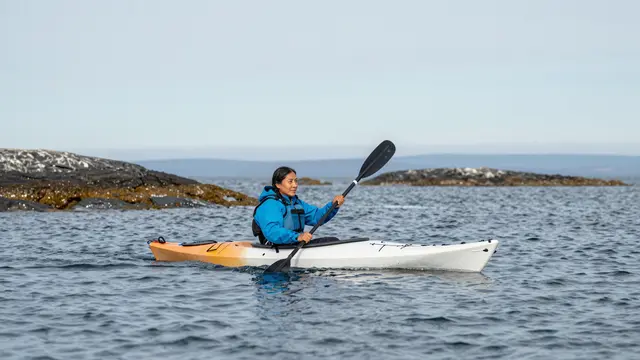 An Indigenous woman in a blue jacket paddles a kayak between small rocky islands on calm open sea