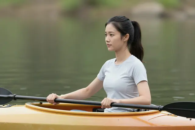Side view of an East Asian woman resting a paddle across her yellow kayak on still green water