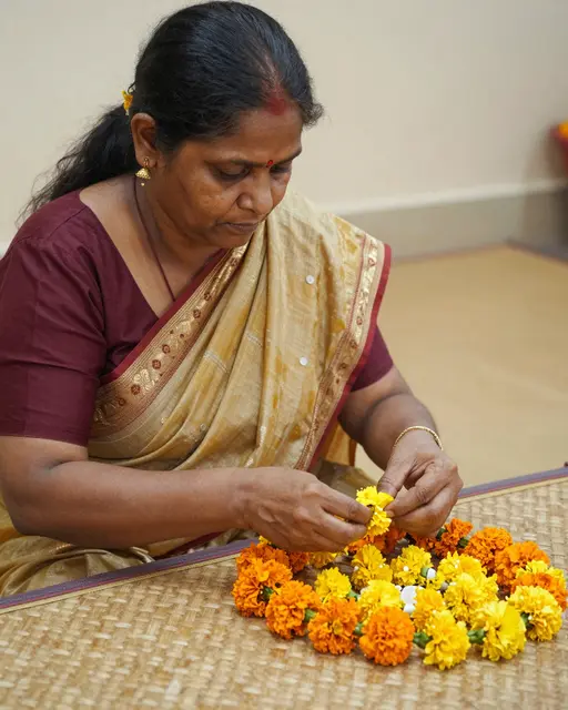 South Asian woman in a sari threads marigold flowers into a garland on a woven mat
