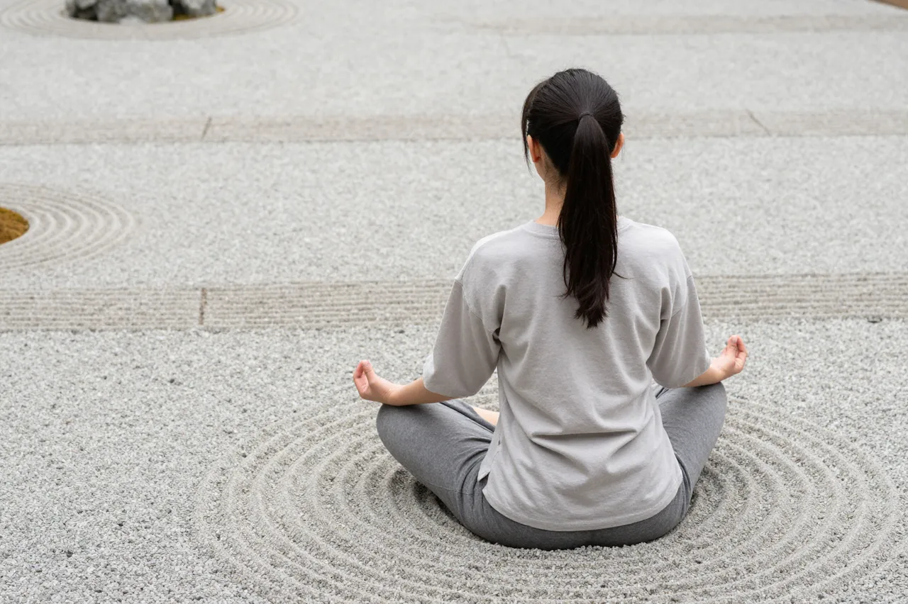 Back view of a woman sitting cross-legged meditating in a raked gravel zen garden