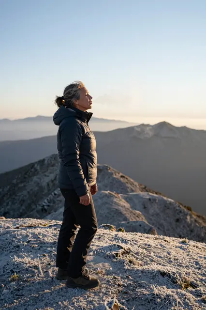 Side view of a middle aged woman standing on a frosty mountain summit, visible breath catching golden dawn light