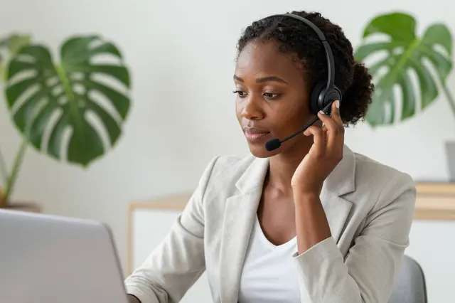 Black woman in her thirties wears a headset while working at a home desk with a laptop and monstera plants