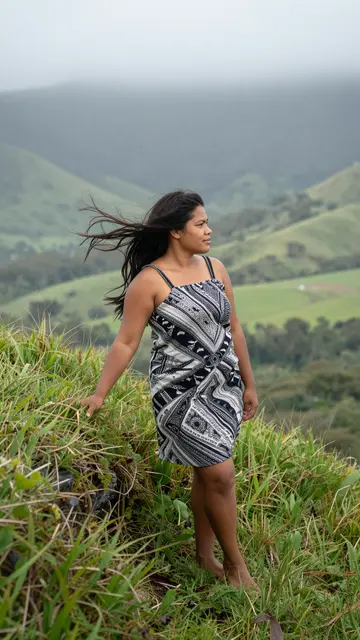 Pacific Islander woman stands barefoot on a grassy ridge as wind blows her hair above a misty valley