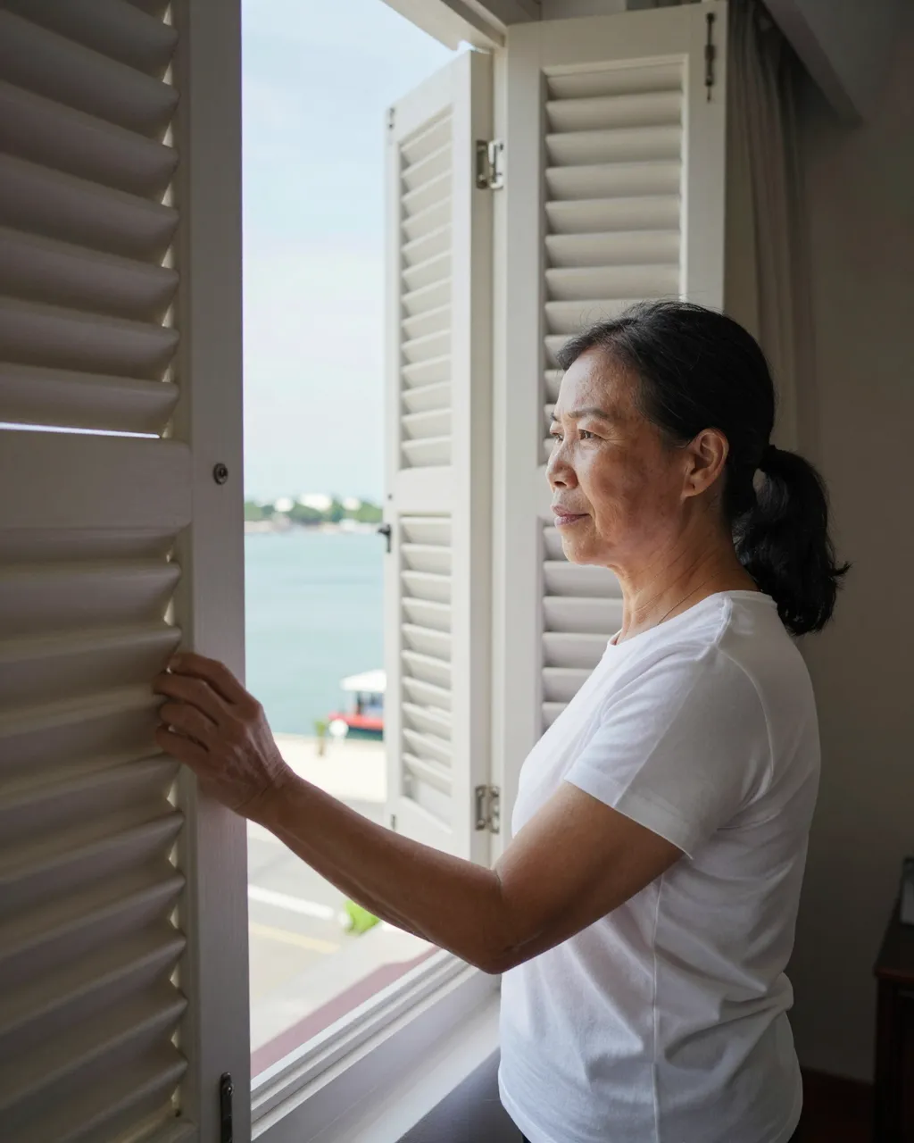 Senior Southeast Asian woman opens window shutters to view a sunny harbor from a hotel room