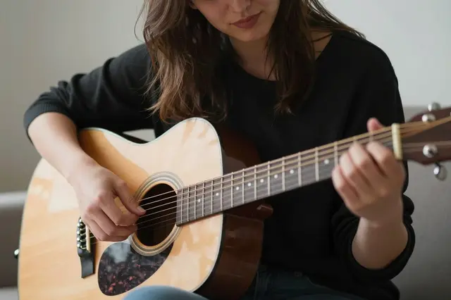 A seated woman plays a light wood acoustic guitar while looking down at the instrument in a home living space