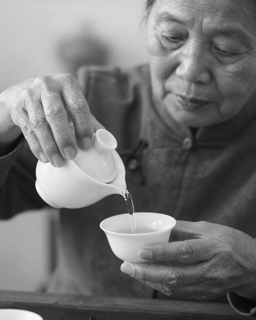 Black and white close-up of an older East Asian woman pouring tea from a small pitcher into a porcelain cup