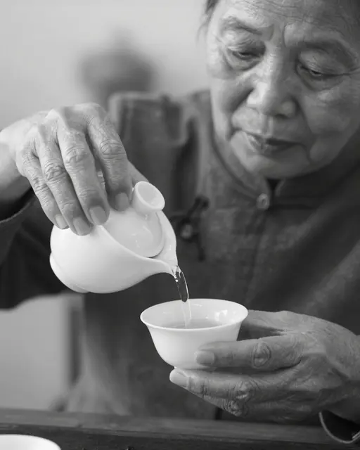 Black and white close-up of an older East Asian woman pouring tea from a small pitcher into a porcelain cup