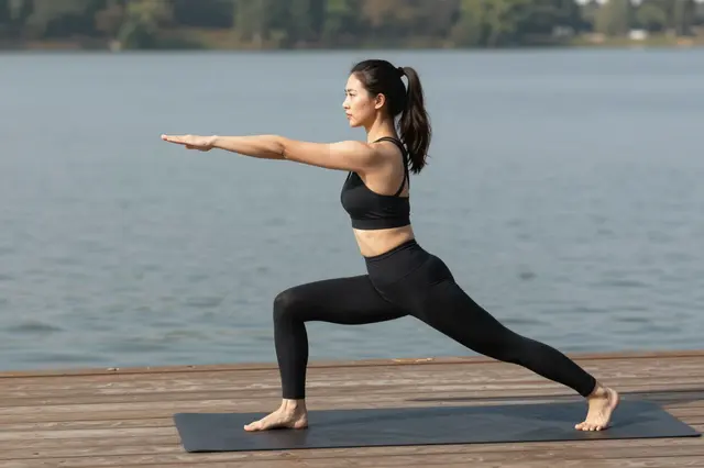 Side view of a woman in a warrior yoga pose standing on a mat on a wooden lakeside deck