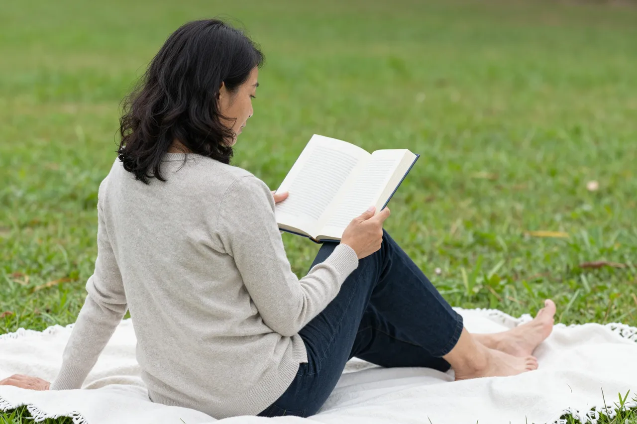 A middle-aged Southeast Asian woman sits barefoot on a blanket reading an open book on park grass