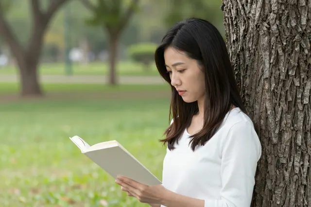 Side view of a woman leaning against a tree while reading an open book in a grassy park