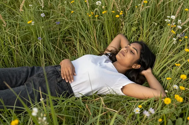 South Asian woman in her twenties lies on her back in tall summer grass with wildflowers, eyes closed and relaxed