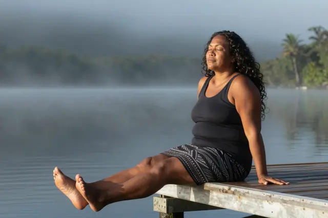 Pacific Islander woman sits on a wooden lake dock with eyes closed during calm misty morning
