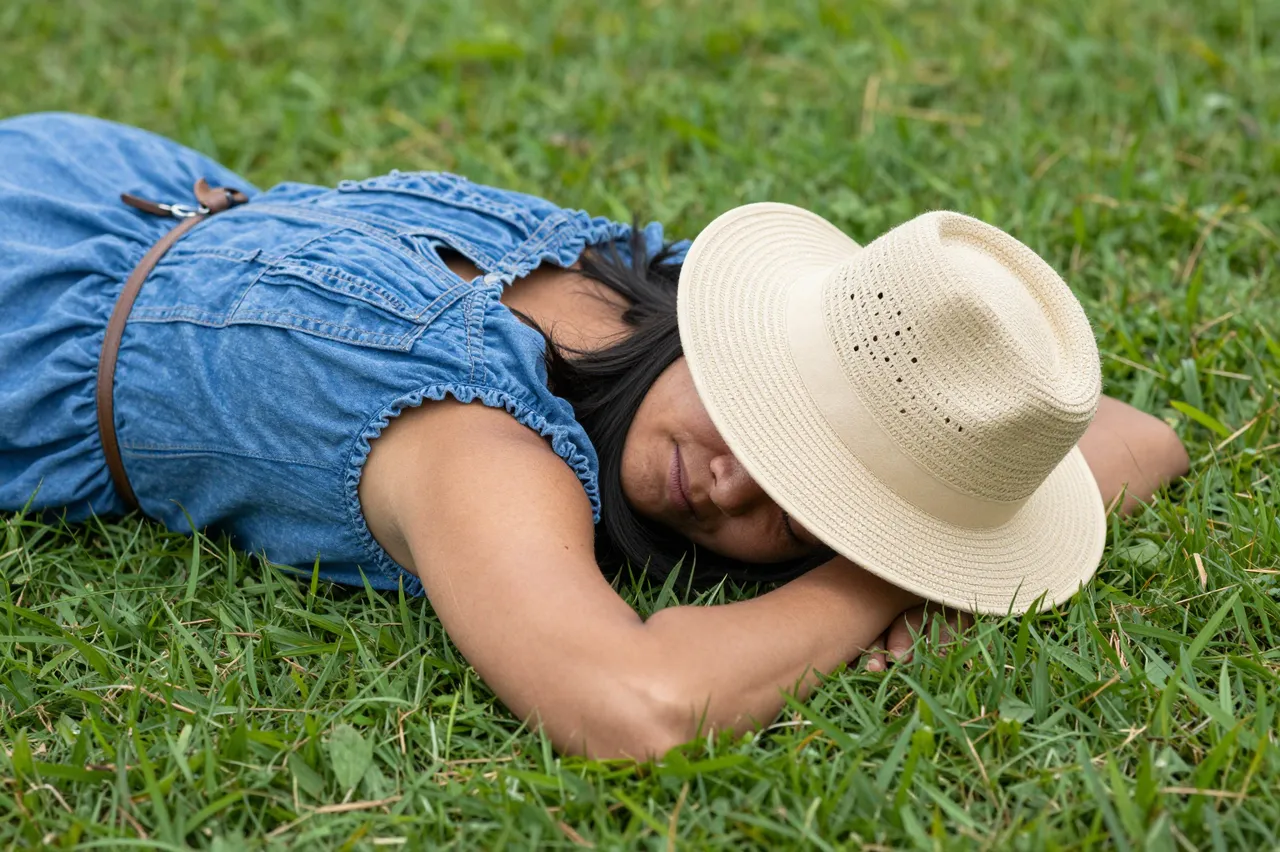 A woman lies on green grass with a straw hat resting over part of her face, relaxing on a summer afternoon