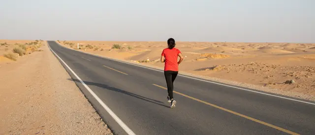 A woman viewed from behind runs alone on a straight paved desert road at dawn casting a long shadow