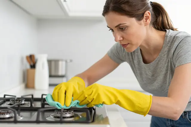 A woman in yellow rubber gloves cleans a gas stovetop with a green cloth in a bright kitchen
