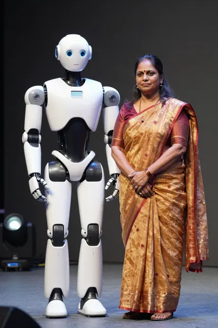 South Asian woman in a gold saree stands beside a white humanoid robot on a lit presentation stage