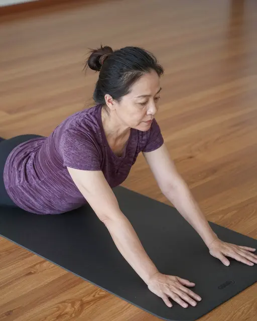 Mature Southeast Asian woman performs a yoga stretch on a black mat over a wooden floor.