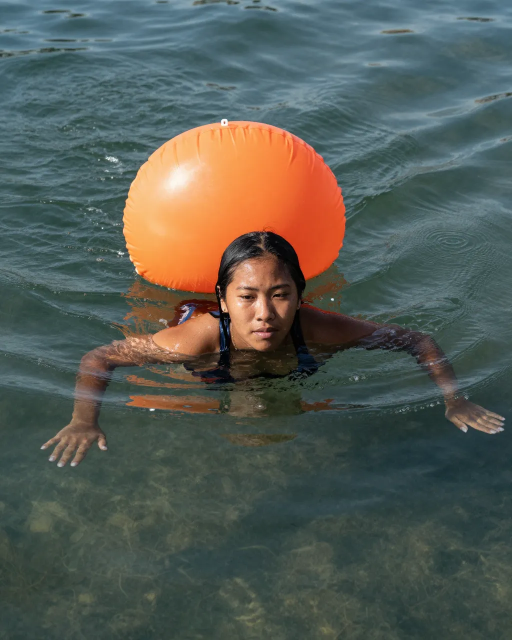A woman swims in clear greenish lake water with a bright orange tow float behind her, viewed from above.