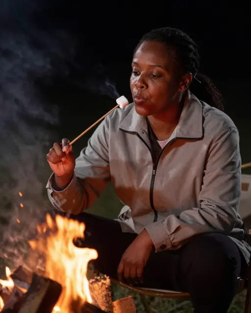 A Black woman in a plain jacket blows on a toasted marshmallow on a stick beside a nighttime campfire