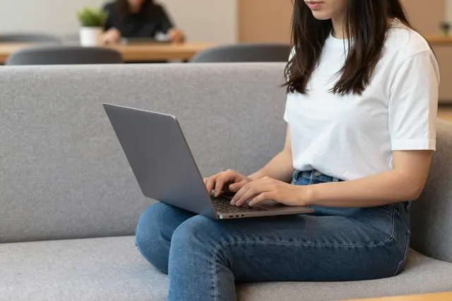 A woman sits cross-legged on a gray sofa typing on a laptop in a coworking lounge, with another blurred person in the