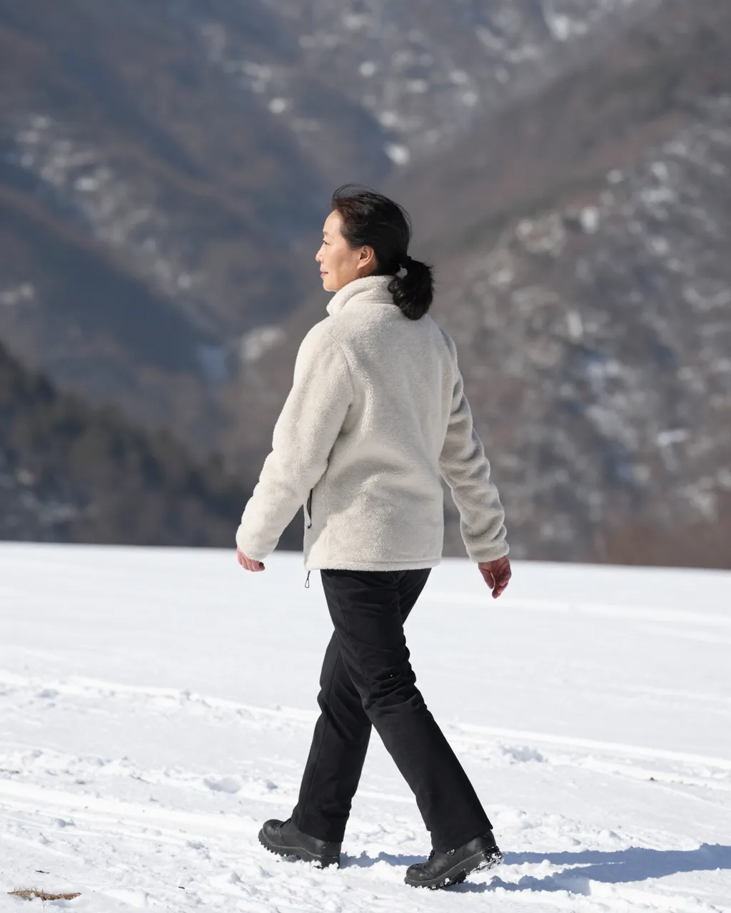 A middle-aged East Asian woman in a cream fleece walks across a sunlit snowy mountain pass with forested peaks in the