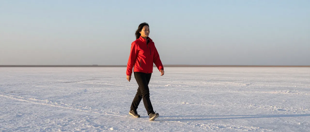 A Southeast Asian woman in a red jacket walks alone across a vast wide salt flat at soft sunrise.