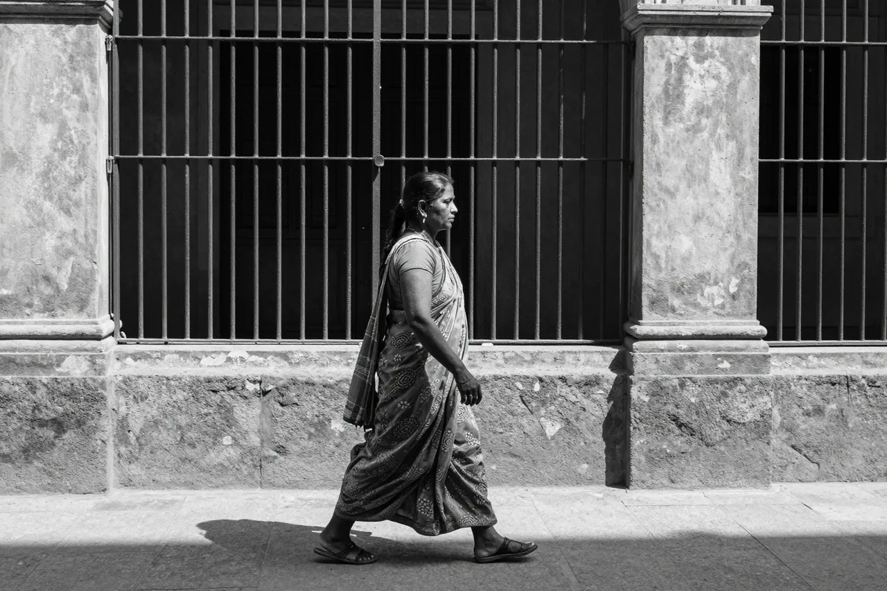A South Asian woman in a sari walks in profile along a sunlit stone arcade behind iron bars in black and white