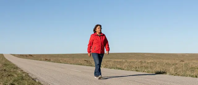 A senior woman in a red jacket walks along an empty paved prairie road under a clear blue sky