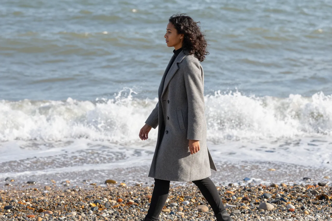 Side view of a mixed-race woman in a gray wool coat walking along a pebbled shore as waves break behind her.