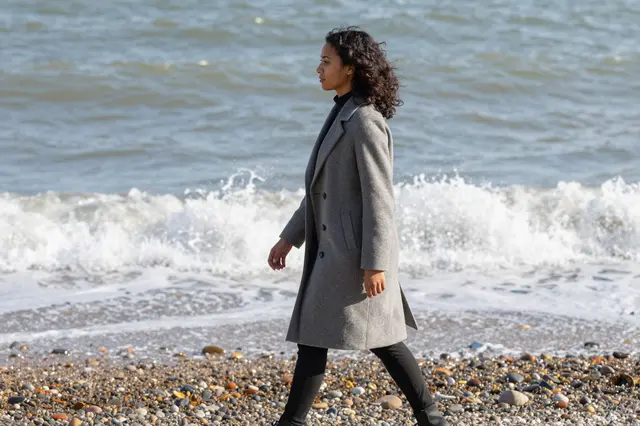 Side view of a mixed-race woman in a gray wool coat walking along a pebbled shore as waves break behind her.