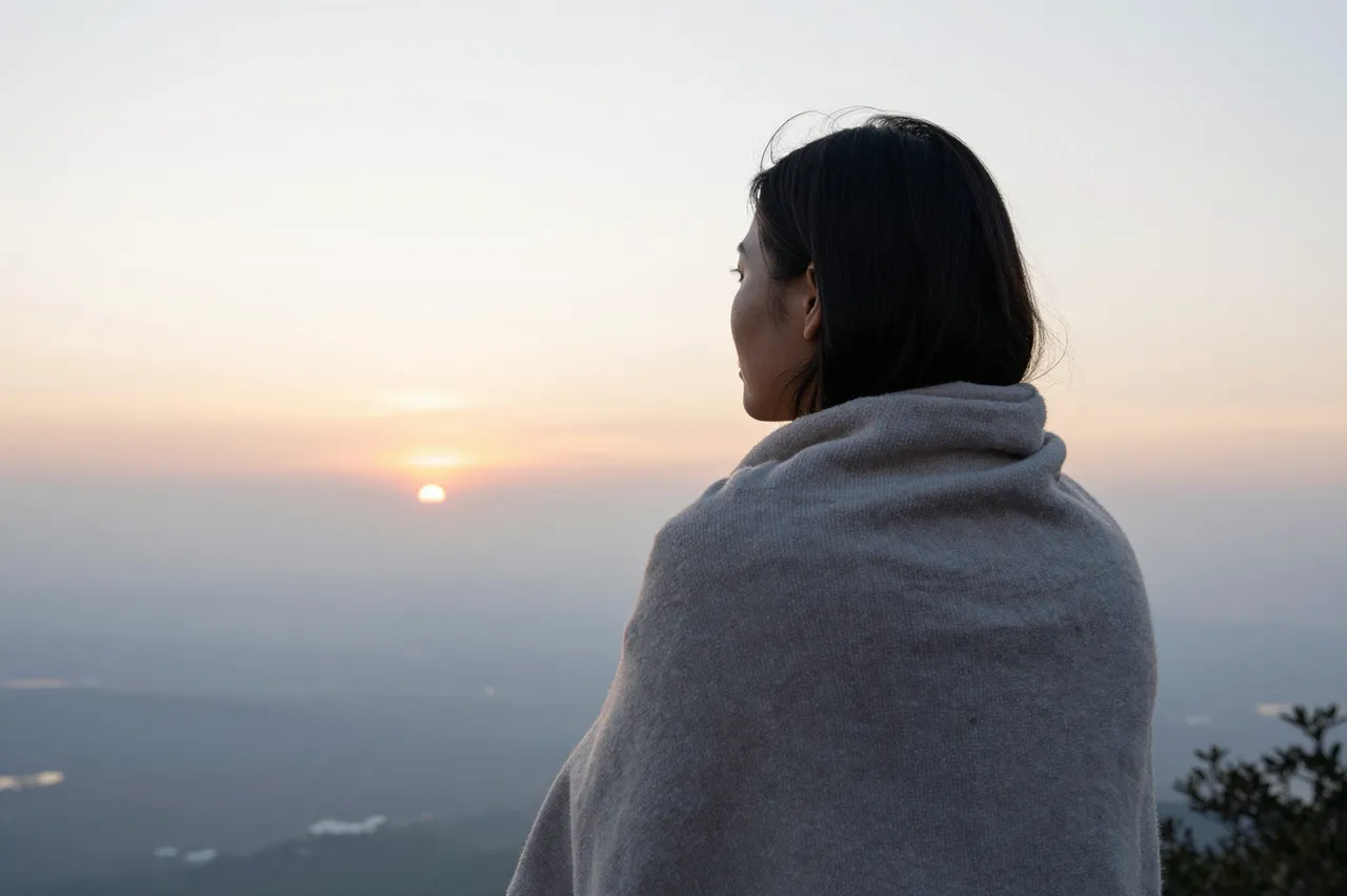 A Southeast Asian woman wrapped in a blanket looks out at sunrise from a hilltop overlook