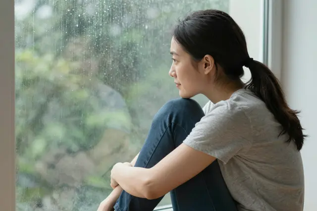 A side view of an adult woman hugging her knees while sitting beside a rain-streaked window