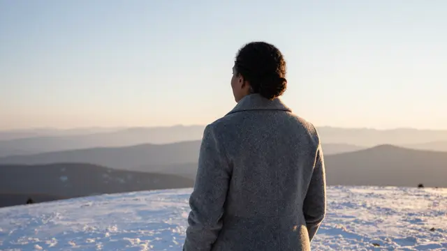 A woman in a wool coat stands from behind on a snowy hilltop watching a pale golden dawn