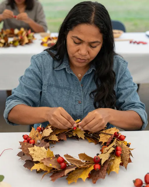 Pacific Islander woman crafts a dried autumn leaf and rose hip wreath at an outdoor craft table