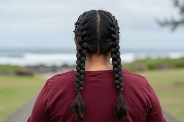 Back view of a woman with two long feed-in braids standing on a seaside path under overcast skies