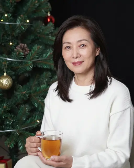 East Asian woman in a white sweater holds a mug of tea while seated next to a decorated Christmas tree at night
