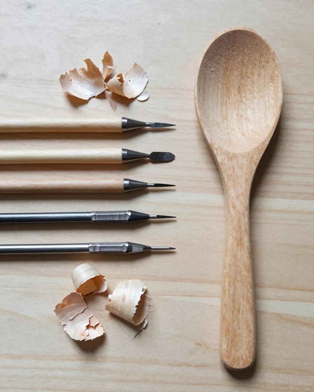 Top-down flat lay of carving tools, pale wood shavings, and a half-finished wooden spoon on a light workbench