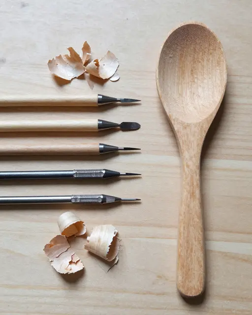 Top-down flat lay of carving tools, pale wood shavings, and a half-finished wooden spoon on a light workbench