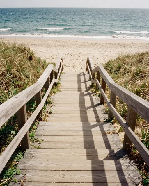 Weathered wooden beach stairs lead down through grassy dunes to a sandy shore and rolling ocean waves.