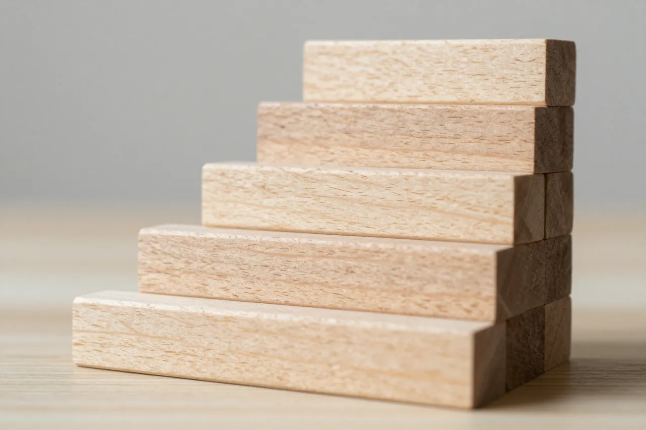 Stacked wooden blocks forming ascending stairs rest on a light wood table against a soft gray background