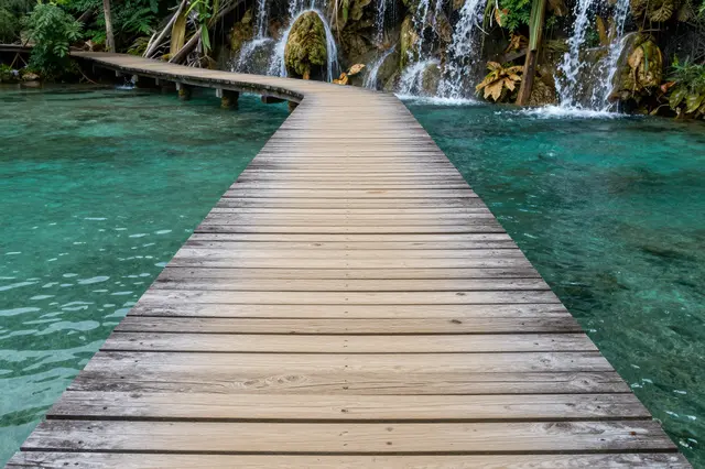A curved wooden walkway stretches over clear turquoise water toward small cascading waterfalls and tropical foliage.