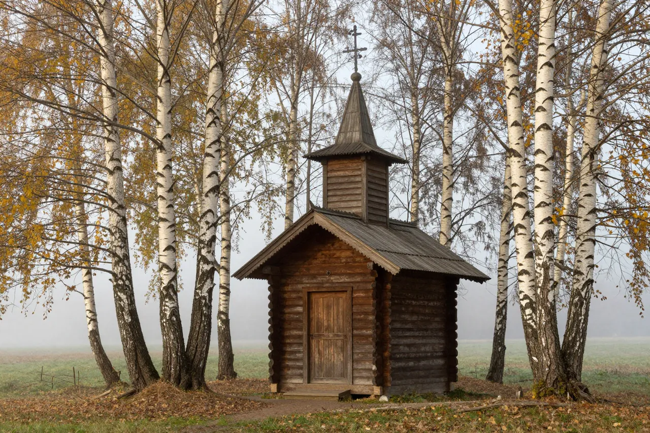 Small log wooden chapel stands between autumn birch trees in soft morning fog