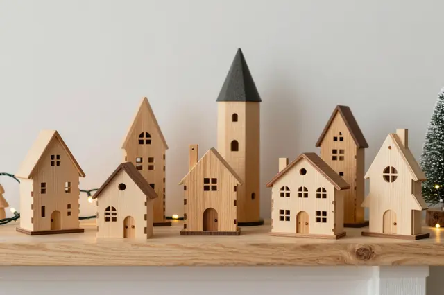Handcrafted wooden Christmas village figurines arranged on a light wood mantel with warm fairy lights and a small decorative