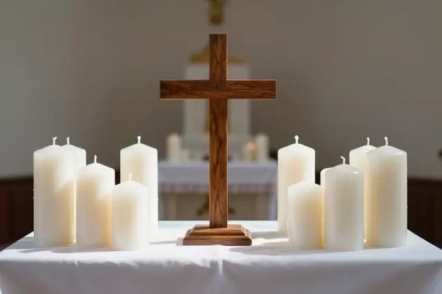 Plain wooden cross surrounded by unlit white pillar candles on a sunlit chapel altar