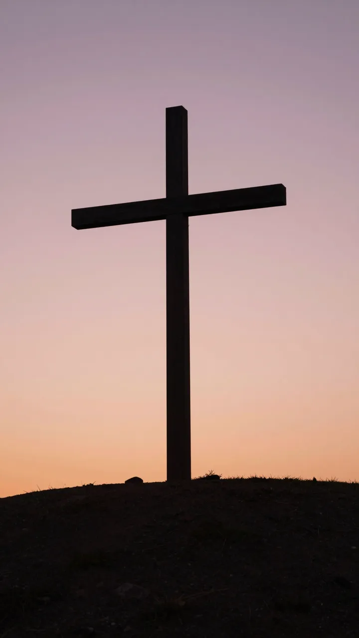 Dark wooden cross silhouetted on a hill against a soft pink and orange gradient dawn sky