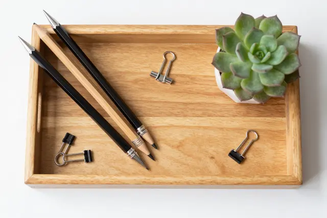Top view of a wooden desk tray holding pencils, binder clips, and a small potted succulent on a white surface