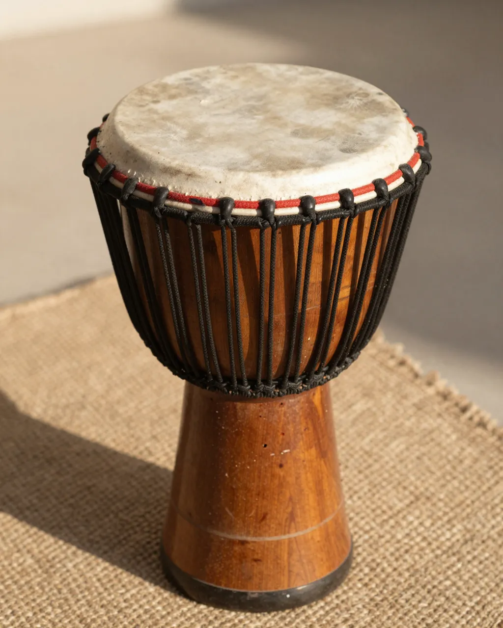 A wooden djembe drum with a natural goatskin head sits on a woven textile in warm afternoon sunlight.