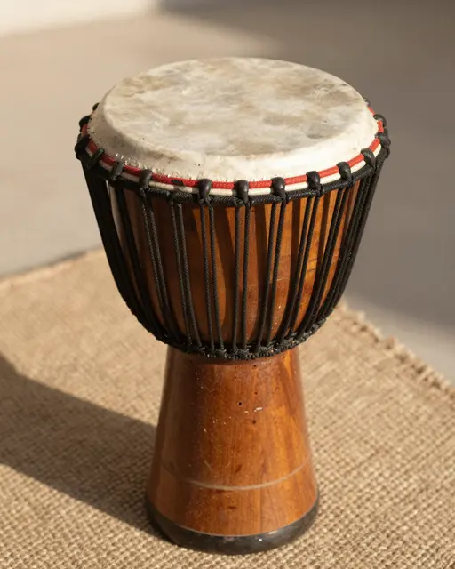 A wooden djembe drum with a natural goatskin head sits on a woven textile in warm afternoon sunlight.