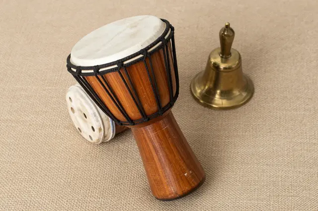 Top view of wooden hand drums and a brass hand bell resting on a woven tan fabric surface