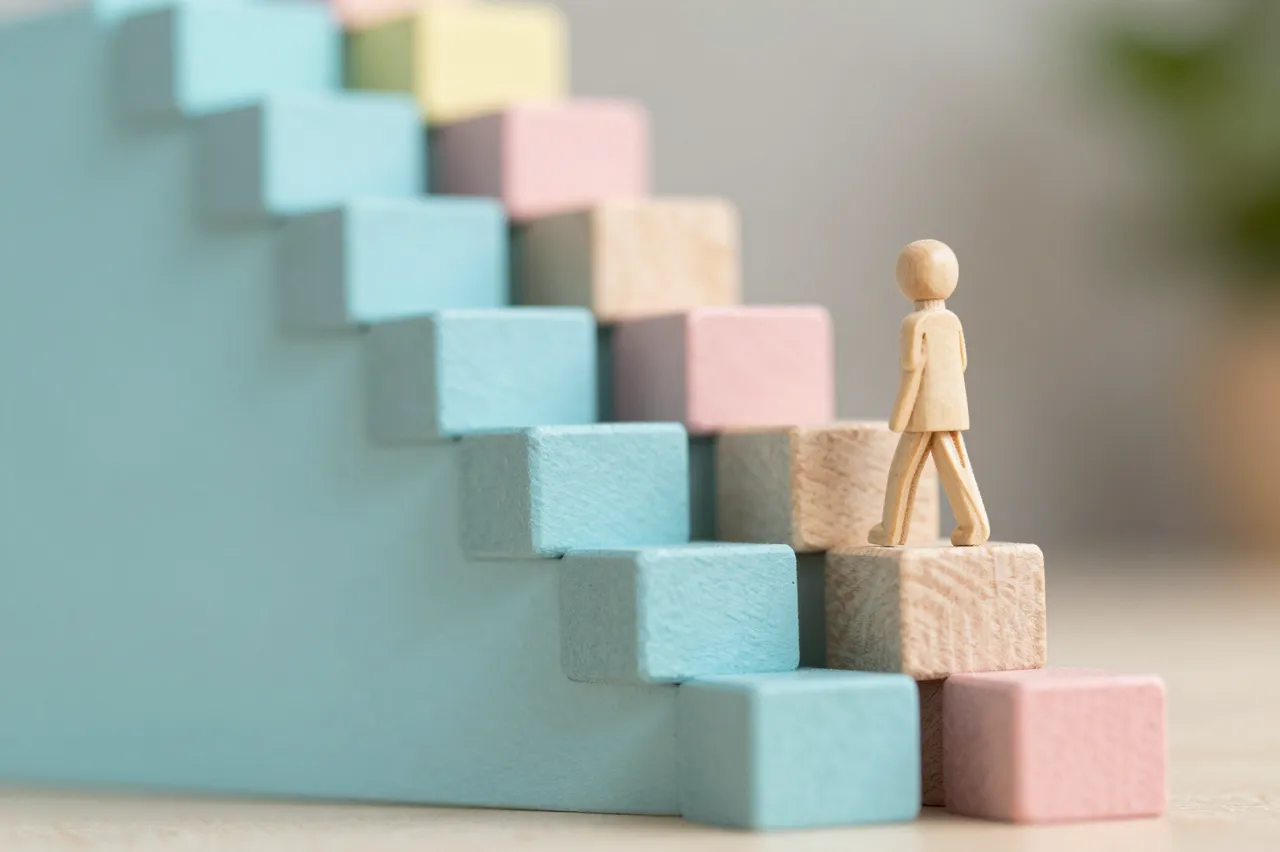 A small wooden figurine stands on pastel wooden block stairs representing career growth